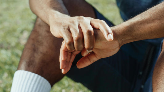 Portraitaufnahme im Gras, Person sitzend, breitbeinig mit weißen Tennissocken, kurzer Hose. Fokus liegt auf Händen: die eine hält die andere  locker umschlungen.